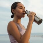 A woman drinks from a metal bottle on a sunny day by the beach, wearing a sports bra.