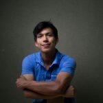 Young Hispanic man smiling confidently in a blue shirt against a dark background.