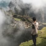 A man stands on a cliff edge overlooking the ancient ruins of Machu Picchu enveloped in mist.